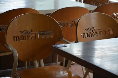 A set of wooden chairs with cut-out lettering on the backrests can be seen. The chairs are arranged around wooden tables, suggesting a cafe or restaurant setting. The lighting is soft and natural, creating subtle reflections on the surfaces.