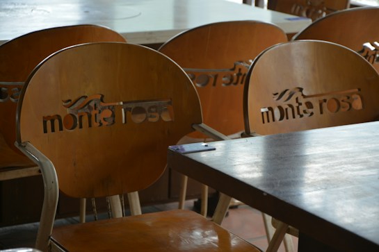 A set of wooden chairs with cut-out lettering on the backrests can be seen. The chairs are arranged around wooden tables, suggesting a cafe or restaurant setting. The lighting is soft and natural, creating subtle reflections on the surfaces.