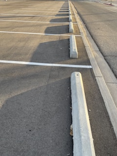 An empty parking lot with lined parking spaces, concrete wheel stops, and shadows cast by the setting sun along the asphalt surface.