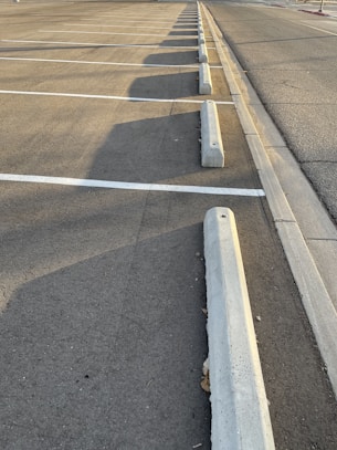 An empty parking lot with lined parking spaces, concrete wheel stops, and shadows cast by the setting sun along the asphalt surface.