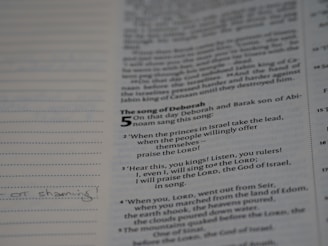 An open book displaying a page with text from 'The Song of Deborah' in a biblical style, featuring verses and numbers. The text is printed in black on an off-white background. There are also lined margins for notes with some handwritten text visible.