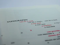 A close-up view of a printed page featuring the word 'TYPOGRAPHY' in bold black letters, followed by several phrases in varying font sizes and colors. The text is creatively arranged with alternating red and black fonts, primarily focusing on the functional and playful aspects of typography.