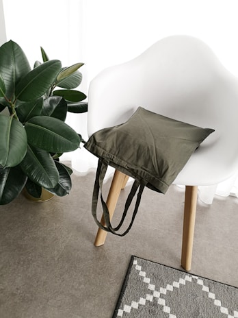 A white modern chair with wooden legs, holding an olive green tote bag. Beside the chair, a lush green potted plant with broad leaves adds a touch of nature. On the floor, a textured gray carpet with a white geometric pattern complements the minimalist setting.