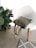 A white modern chair with wooden legs, holding an olive green tote bag. Beside the chair, a lush green potted plant with broad leaves adds a touch of nature. On the floor, a textured gray carpet with a white geometric pattern complements the minimalist setting.