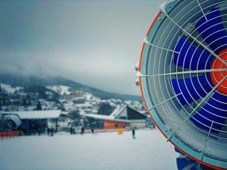 A large snow-making machine is prominently displayed on the right, featuring vivid blue and orange blades encased in a metal grate. The background consists of a snow-covered village with several buildings and a few people walking or skiing in the distance. Snow blankets the ground and creates a serene winter atmosphere.