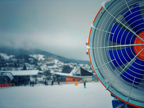 A large snow-making machine is prominently displayed on the right, featuring vivid blue and orange blades encased in a metal grate. The background consists of a snow-covered village with several buildings and a few people walking or skiing in the distance. Snow blankets the ground and creates a serene winter atmosphere.