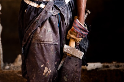 A close-up view of a person holding a hammer by the handle, wearing worn-out, paint-spattered work pants. The individual is wearing a glove on one hand, gripping the handle of a tool, possibly giving the impression of manual labor. The background is blurred and dark, emphasizing the focus on the lower part of the person and the tool they are holding.