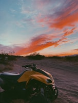 A yellow ATV parked on a sandy path in a desert landscape during sunset. The sky is filled with vibrant orange and pink clouds, contrasting with the blue hues. Sparse desert vegetation surrounds the path, with silhouettes of low hills in the distance.