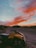 A yellow ATV parked on a sandy path in a desert landscape during sunset. The sky is filled with vibrant orange and pink clouds, contrasting with the blue hues. Sparse desert vegetation surrounds the path, with silhouettes of low hills in the distance.