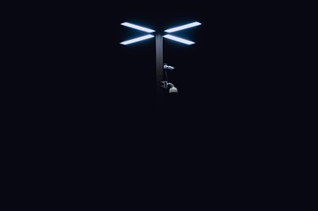 A modern streetlight with dual LED panels glows brightly against a dark background. Below the lights, there are various security cameras attached to the pole.