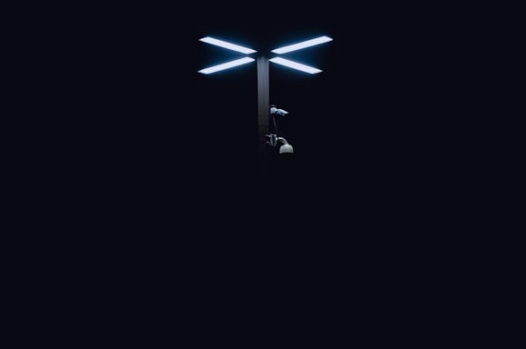 A modern streetlight with dual LED panels glows brightly against a dark background. Below the lights, there are various security cameras attached to the pole.