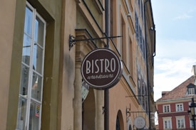 A street view with a sign for a bistro named Warszawa hanging from a building. The architecture features traditional, colorful European-style buildings with windows and decorative elements along the facade. The sky is clear and light blue.