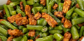 A close-up shot of a dish featuring crispy tempeh pieces mixed with fresh, vibrant green beans. The tempeh has a rich brown color with a hearty texture, while the green beans are cut into short segments and appear glossy from being saut&eacute;ed or stir-fried.