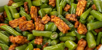 A close-up shot of a dish featuring crispy tempeh pieces mixed with fresh, vibrant green beans. The tempeh has a rich brown color with a hearty texture, while the green beans are cut into short segments and appear glossy from being sautéed or stir-fried.