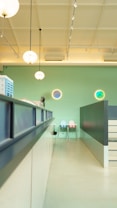 A modern, brightly lit interior space with a high ceiling and pendant lights hanging down. The walls are painted in a light green color, and there are circular decorative elements on the wall with blue and green tones. In the background, there are two pastel-colored chairs against the wall, one in light blue and the other in light pink. A counter or desk runs along the left side with some equipment placed on it. The overall ambiance appears clean and minimalistic.