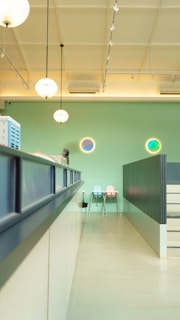 A modern, brightly lit interior space with a high ceiling and pendant lights hanging down. The walls are painted in a light green color, and there are circular decorative elements on the wall with blue and green tones. In the background, there are two pastel-colored chairs against the wall, one in light blue and the other in light pink. A counter or desk runs along the left side with some equipment placed on it. The overall ambiance appears clean and minimalistic.