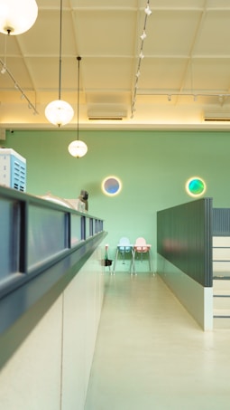 A modern, brightly lit interior space with a high ceiling and pendant lights hanging down. The walls are painted in a light green color, and there are circular decorative elements on the wall with blue and green tones. In the background, there are two pastel-colored chairs against the wall, one in light blue and the other in light pink. A counter or desk runs along the left side with some equipment placed on it. The overall ambiance appears clean and minimalistic.