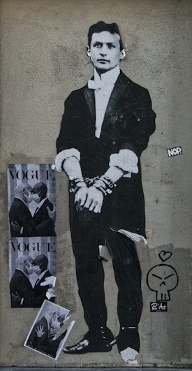 A black-and-white street art piece features a figure in a formal suit, depicted in a vintage style with their hands in chains. To the left, two posters show mirror image couples in masks embracing with the word 'VOGUE' printed across. Below, a smaller torn image shows part of a couple in a similar pose. A small cartoonish skull with a heart above it and the signature 'Pen'Art' is visible next to the figure.