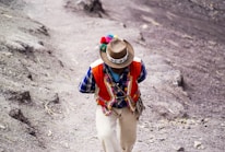 A person wearing traditional Andean clothing is walking on a dirt path. Their attire includes a colorful vest adorned with patterns, a checkered shirt, and a hat with decorative elements. The surrounding landscape is earthy and rugged.