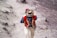 A person wearing traditional Andean clothing is walking on a dirt path. Their attire includes a colorful vest adorned with patterns, a checkered shirt, and a hat with decorative elements. The surrounding landscape is earthy and rugged.