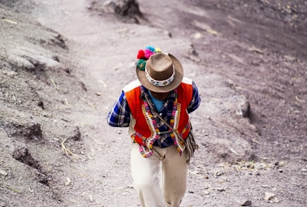 A person wearing traditional Andean clothing is walking on a dirt path. Their attire includes a colorful vest adorned with patterns, a checkered shirt, and a hat with decorative elements. The surrounding landscape is earthy and rugged.
