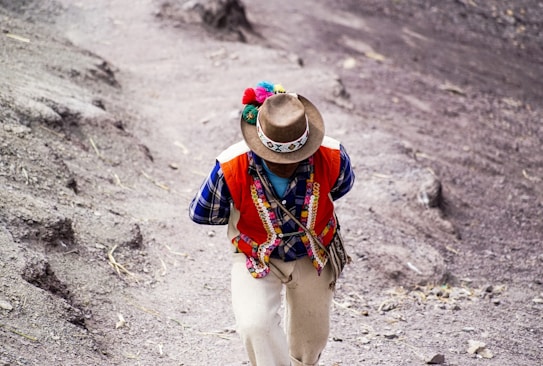 A person wearing traditional Andean clothing is walking on a dirt path. Their attire includes a colorful vest adorned with patterns, a checkered shirt, and a hat with decorative elements. The surrounding landscape is earthy and rugged.