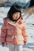A child wearing a red and white checkered coat stands in the snow. The child has straight black hair and appears to be thoughtful or contemplative. In the background, a blurred figure of a giraffe is visible.