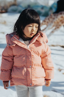 A child wearing a red and white checkered coat stands in the snow. The child has straight black hair and appears to be thoughtful or contemplative. In the background, a blurred figure of a giraffe is visible.