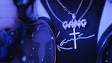 A close-up of a person wearing a shiny chain with a cross pendant and a separate pendant spelling 'GANG'. The person is also wearing a dark shirt with a stylized signature text and a jacket featuring an artistic pattern, all under a blue-tinted lighting.