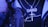 A close-up of a person wearing a shiny chain with a cross pendant and a separate pendant spelling 'GANG'. The person is also wearing a dark shirt with a stylized signature text and a jacket featuring an artistic pattern, all under a blue-tinted lighting.