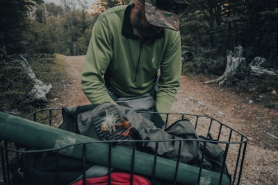 A person wearing a camouflage cap and green long-sleeve shirt is leaning over a metal basket filled with camping or fishing gear. The setting is a wooded outdoor area with a dirt path, surrounded by trees and fallen logs.