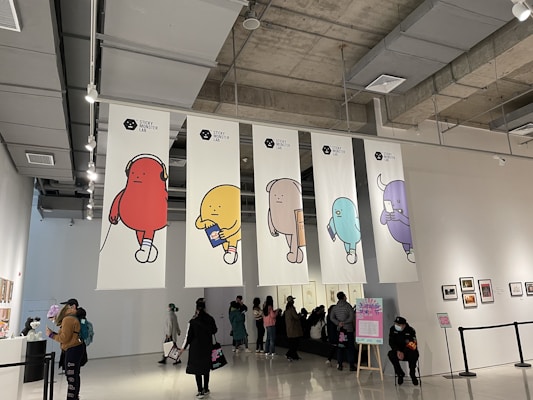 A gallery space with high ceilings and exposed ductwork has large banners depicting colorful cartoon-like characters. People are casually walking around, viewing artwork displayed on the walls. A person is sitting in front of a sign with text in the foreground.