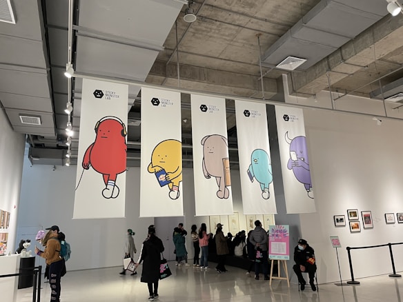 A gallery space with high ceilings and exposed ductwork has large banners depicting colorful cartoon-like characters. People are casually walking around, viewing artwork displayed on the walls. A person is sitting in front of a sign with text in the foreground.