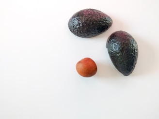 Two whole avocados and one avocado pit are placed against a plain white background. The avocados are dark green with a bumpy texture, and the seed is brown and round.