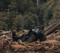 A person in outdoor attire reclines against an all-terrain vehicle situated amidst a rocky, forested landscape. The scene conveys a rugged, adventurous atmosphere with dense greenery and uneven terrain.
