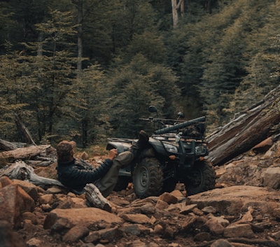 A person in outdoor attire reclines against an all-terrain vehicle situated amidst a rocky, forested landscape. The scene conveys a rugged, adventurous atmosphere with dense greenery and uneven terrain.