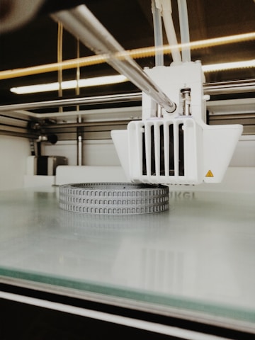 A close-up view of a 3D printer in action, focusing on the extruder head and the partially printed object on the print bed. The object appears to be a circular structure with small, repetitive grooves on its surface.