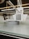 A close-up view of a 3D printer in action, focusing on the extruder head and the partially printed object on the print bed. The object appears to be a circular structure with small, repetitive grooves on its surface.