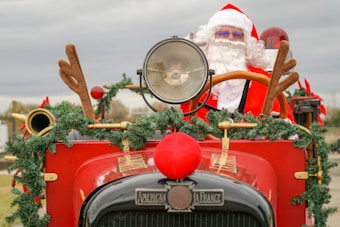 A person dressed as Santa Claus is sitting in a vintage red fire truck decorated with festive garlands and reindeer antlers attached to the vehicle. The scene suggests a holiday-themed setting.