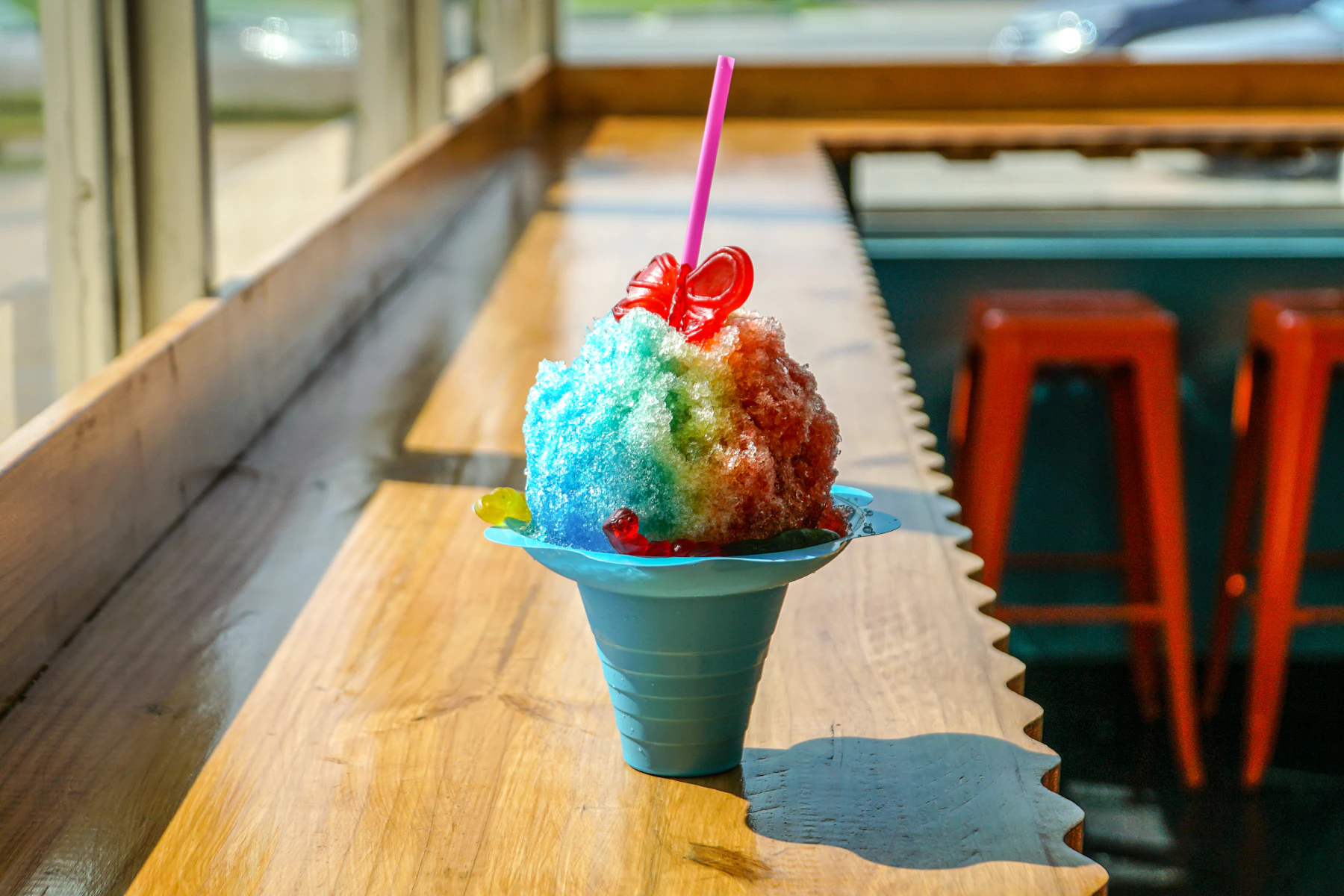 A rainbow snow cone of blue, red and orange syrup in a blue flower-shaped cup on a sunny wooden counter
