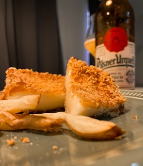 A close-up view of two pieces of breaded and fried cheese served on a plate, accompanied by potato wedges. In the background, there is a bottle of Pilsner Urquell beer with a red circle logo, partially blurred.