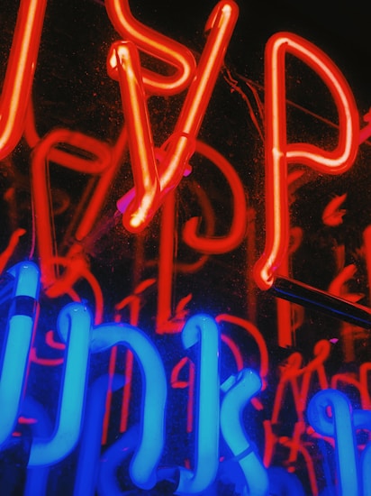 Brightly glowing neon lights in red and blue with a reflective surface creating a vibrant and colorful display. The neon tubes are formed in various shapes and letters, giving an abstract appearance.