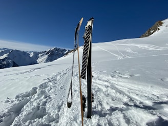Two skis and ski poles are stuck upright in deep snow on a sunny mountain slope. In the background, majestic snow-covered mountains stretch into the distance under a clear blue sky. Multiple ski tracks are visible in the snow, indicating recent activity in a serene alpine environment.