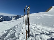 Two skis and ski poles are stuck upright in deep snow on a sunny mountain slope. In the background, majestic snow-covered mountains stretch into the distance under a clear blue sky. Multiple ski tracks are visible in the snow, indicating recent activity in a serene alpine environment.