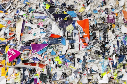 A chaotic assemblage of torn and overlapping colorful paper pieces, stapled haphazardly to a bulletin board or wall. The various layers show fragments of text and images, with sharp edges and jagged shapes creating a textured and dynamic surface.