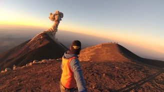 A person with an orange vest and a cap is taking a selfie on a mountain ridge with a smoking volcano in the background. The sun is setting, casting a warm light across the landscape. In the distance, a group of people can be seen standing on another ridge, observing the scene.