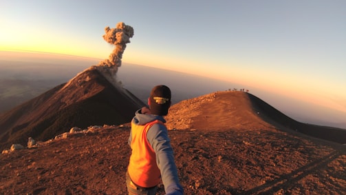 A person with an orange vest and a cap is taking a selfie on a mountain ridge with a smoking volcano in the background. The sun is setting, casting a warm light across the landscape. In the distance, a group of people can be seen standing on another ridge, observing the scene.