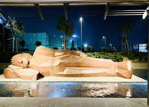 A large, geometric sculpture of a reclining figure is positioned on a platform adjacent to a reflective pool. The sculpture appears to be made from a faceted, orange-brown material. Surrounding the sculpture are neatly trimmed hedges, small trees, and ambient lighting set against a dark evening sky.