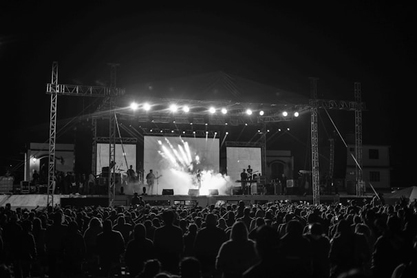 A live music concert with a large crowd gathered in front of an illuminated stage. Musicians are performing under bright lighting, with smoke effects adding to the atmosphere. The scene is captured in black and white, emphasizing contrasts and shadows.