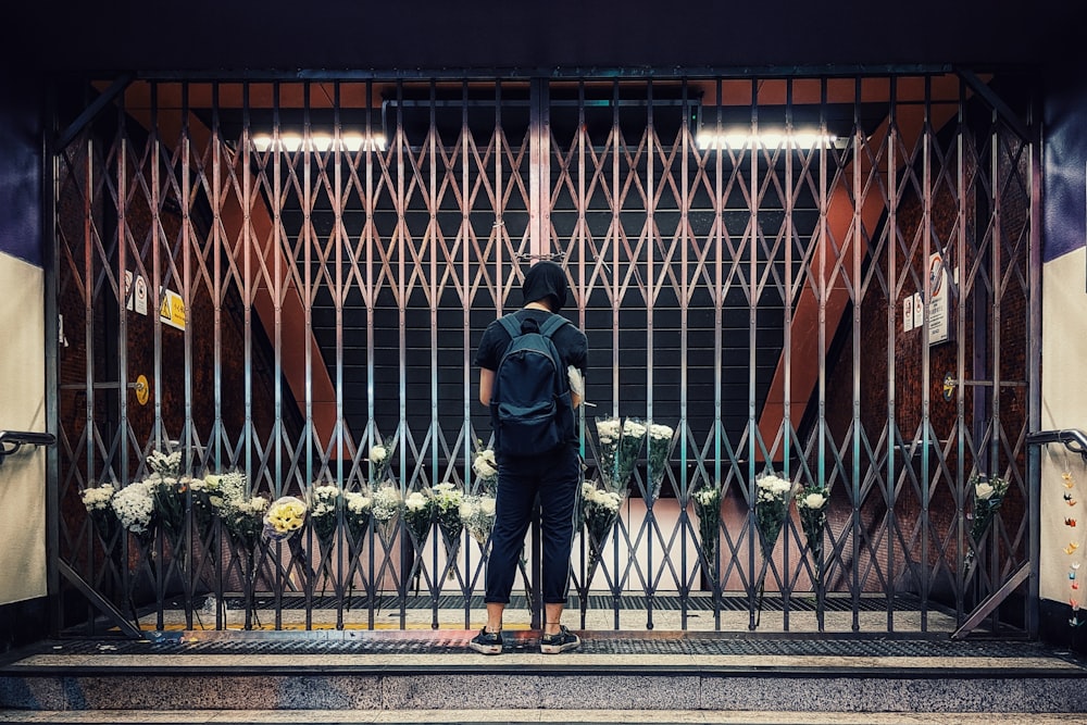 Hombre con chaqueta negra y jeans de mezclilla azules de pie frente a una valla de metal negro